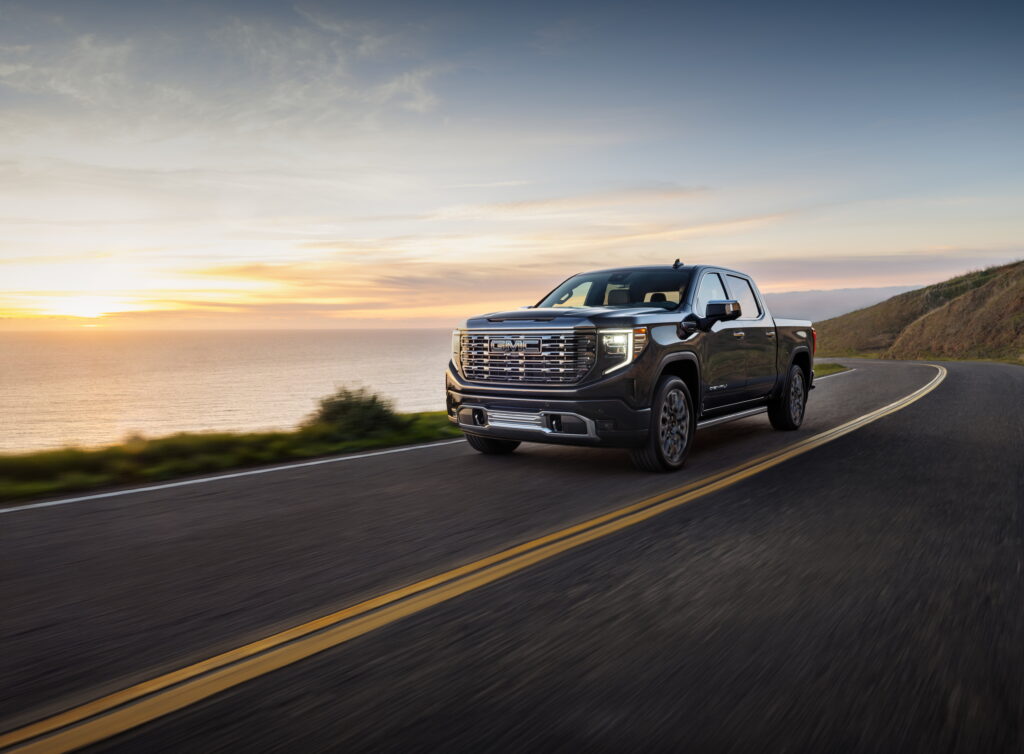 2024 GMC Sierra Denali Ultimate 1500 Crew Cab Short Box in Titanium Rush Metallic with 7-spoke ultra-bright machined wheels; 3/4 driver-side front view in motion next to water.